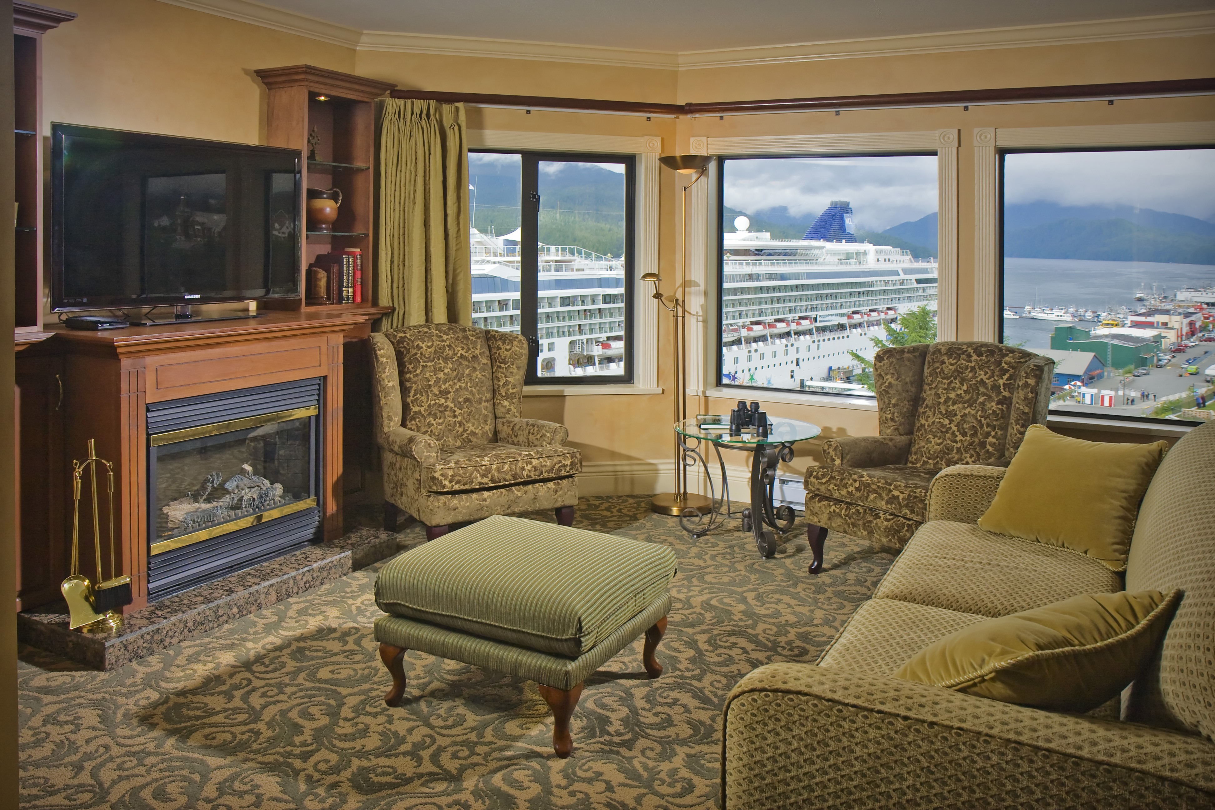 Hotel room with a cozy seating area, including two armchairs, a sofa, and a coffee table. The large windows offer a view of a cruise ship and a port area outside.